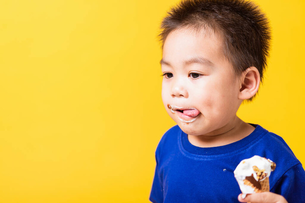 Happy portrait Asian child or kid cute little boy attractive laugh smile playing holds and eating sweet chocolate ice cream waffle cone, studio shot isolated on yellow background, summer concept - Foto, Imagen