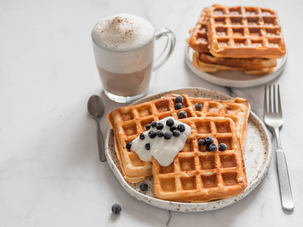 Delicious homemade baked belgian waffles with greek yogurt, blueberries and cappuccino on white marble background. Perfect breakfast with copy space for text or design - Φωτογραφία, εικόνα