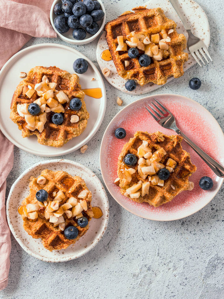 Easy healthy gluten free oat waffles with copy space. Plates with appetizing homemade waffles with oat flour decorated blueberries, bananas and honey on gray background. Vertical. Top view or flat lay - Фото, изображение