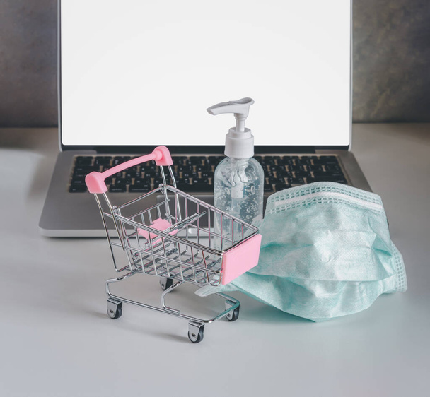 Empty shopping cart, medical face mask, hand alcohol gel and laptop. Online. Stay home, healthcare and shopping concept during quarantine period - Фото, изображение