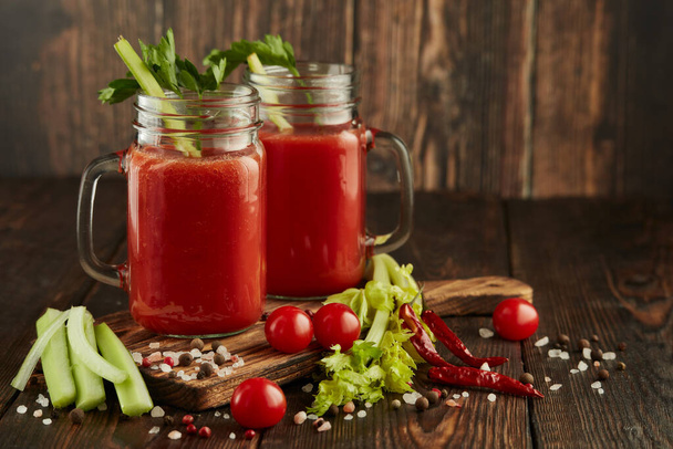 Two glasses with fresh tomato juice, celery, parsley, spices and ripe tomatoes on dark brown wooden background with copy space. Healthy beverage, vegetable juice - Photo, Image