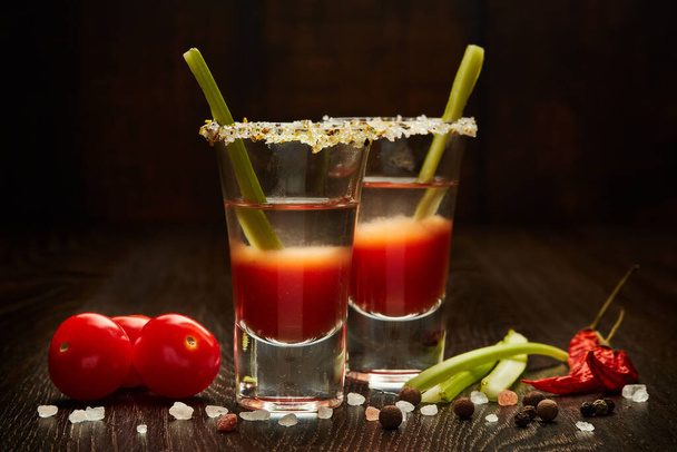 Two shot glasses with cocktail Bloody Mary, celery, spices and ripe tomatoes on dark wooden background - Fotoğraf, Görsel