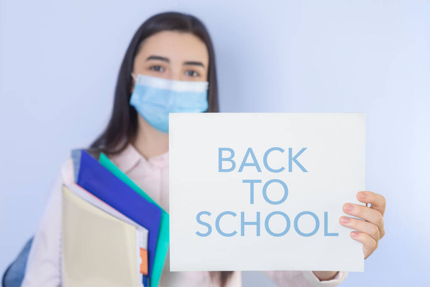 High school girl with mask on her showing back to school message. Student girl ready for school during the coronavirus pandemic. Focus on her hand with paper. - Foto, Bild