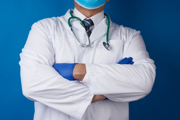 adult male doctor in a white medical coat and blue latex gloves stands on a blue background, his arms crossed on his chest - Valokuva, kuva