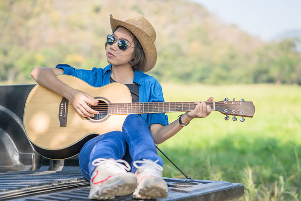 Young asian women short hair wear hat and sunglasses playing guitar ,sit on pickup truck in countryside Thailand - Фото, зображення