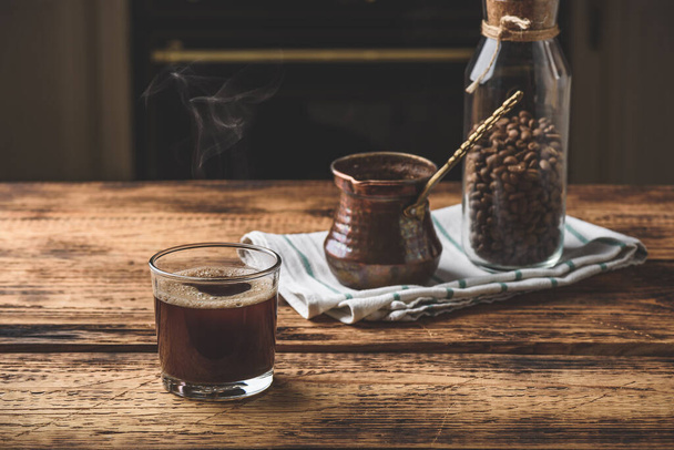 Freshly brewed turkish coffee in drinking glass. Cezve and jar of roasted coffee beans. - Фото, изображение