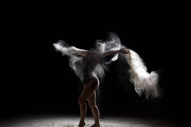 beautiful caucasian woman in a black bodysuit with a sports figure is dancing in a white cloud of flour on a black background - Foto, immagini