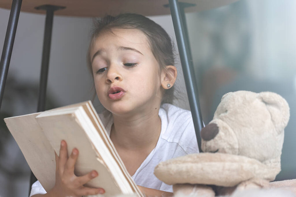 Πορτρέτο ενός χαριτωμένου μικρού κοριτσιού που διαβάζει ένα βιβλίο στο σπίτι, ξαπλωμένη στο πάτωμα με το αγαπημένο της παιχνίδι. - Φωτογραφία, εικόνα