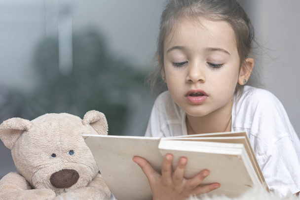 Портрет милой маленькой девочки, читающей книгу дома, лежащей на полу со своей любимой игрушкой. - Фото, изображение