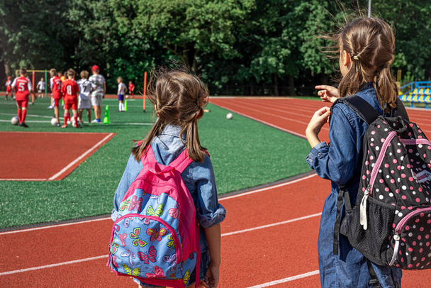 小さな女の子の学校の子供たちはスタジアムでバックパックを着て少年たちがサッカーを見て. - 写真・画像