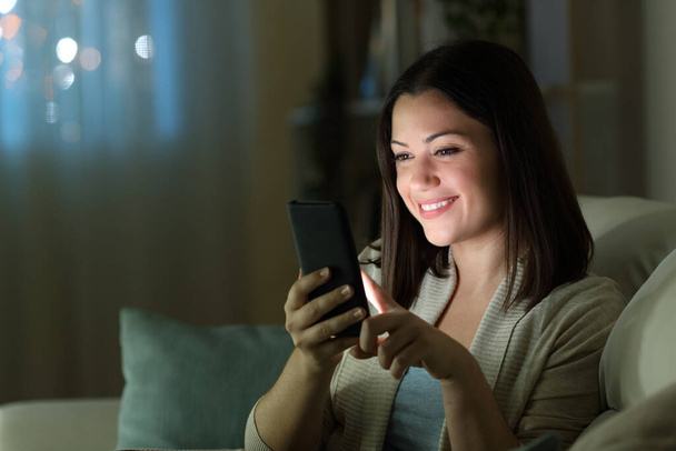 Happy woman using cell phone sititng on a couch in the night at home - Zdjęcie, obraz