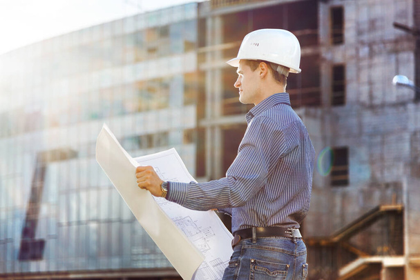 Architect in helmet with blueprints looks at camera in a building site - Photo, Image
