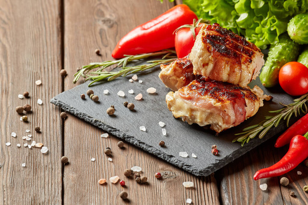 Bacon-wrapped grilled chicken (turkey) fillet with fresh vegetables and spices on stone cutting board on wooden background - Fotoğraf, Görsel