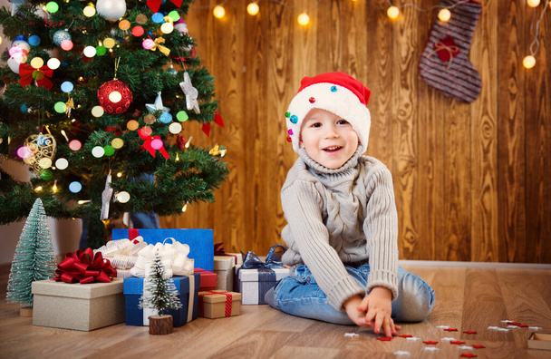 Boy and sitting on the floor with presents near christmas tree. Child in red hat at home in winter - Fotografie, Obrázek
