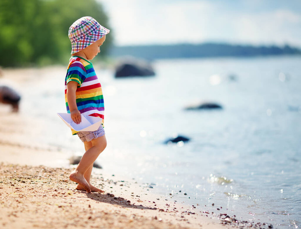two years old boy playing at the beach in hat. Child with a paper ship at sea - Fotoğraf, Görsel