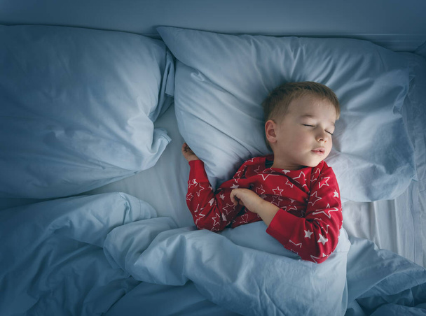 Sleepy boy lying in bed with blue beddings. Tired child in bedroom sleeping. Little kid lying asleep in red pajamas - Foto, afbeelding
