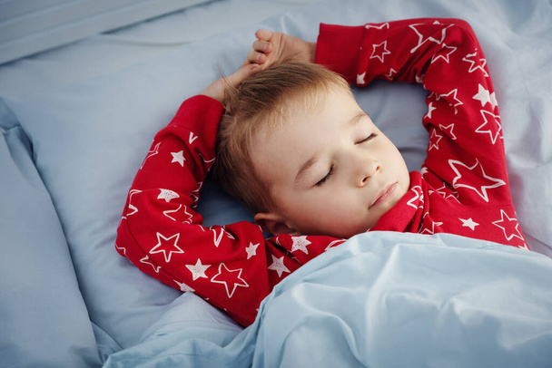 Sleepy boy lying in bed with blue beddings. Tired child in bedroom sleeping. Little kid lying asleep in red pajamas - Valokuva, kuva