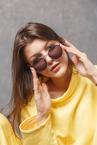 Young woman in yellow sweatshirt and sunglasses on a gray background. - Fotografie, Obrázek