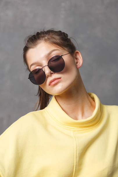 Portrait of young girl weared in yellow sweetshirt in sunglasses on a gray background. - Fotoğraf, Görsel
