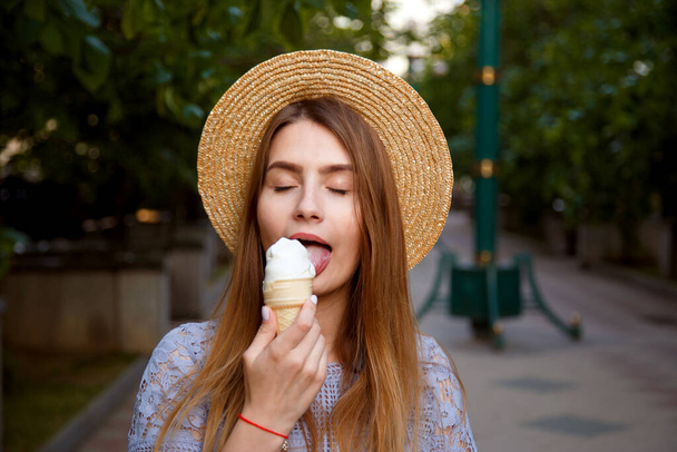 Young caucasian woman in hat enjoying ice cream outdoors in summer. Girl close her eyes in pleasure - Фото, изображение