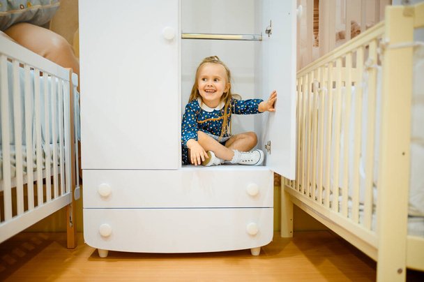 Smiling little girl sitting in wardrobe near baby bed in kid's store. Adorable child at the showcase in children's shop, happy childhood, gaughter makes a purchase in shop - Foto, Bild