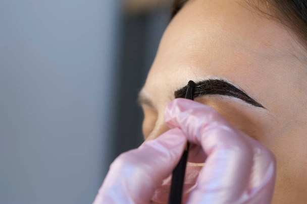 Cosmetologist is painting womans eyebrows in cosmetology clinic, closeup view. - 写真・画像