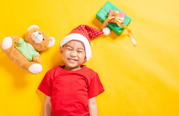 Asian little cute child boy smile, Top view of a kid dressed in red Santa Claus hat the concept of holiday Christmas Xmas day or Happy new year, isolated lying on yellow background - Foto, imagen