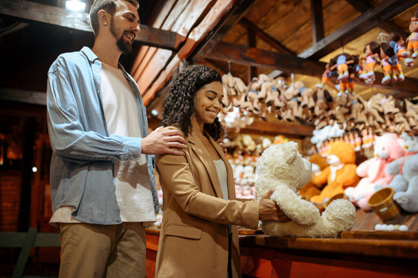 Love couple wins soft toy in amusement park souvenir shop. Man and woman relax outdoors, fairground on background. Family leisures on carousels, entertainment theme - Fotó, kép