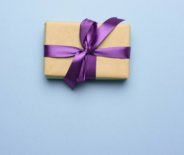 box wrapped in brown paper and tied with a purple silk ribbon with a bow, gift on a blue background, top view - Фото, изображение