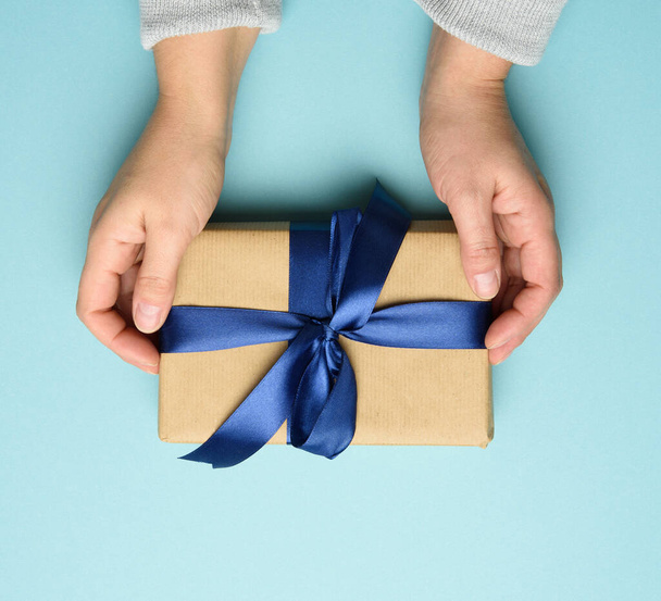 two female hands in a sweater hold a square gift box with a blue bow on a blue background, top view, festive backdrop - Foto, afbeelding