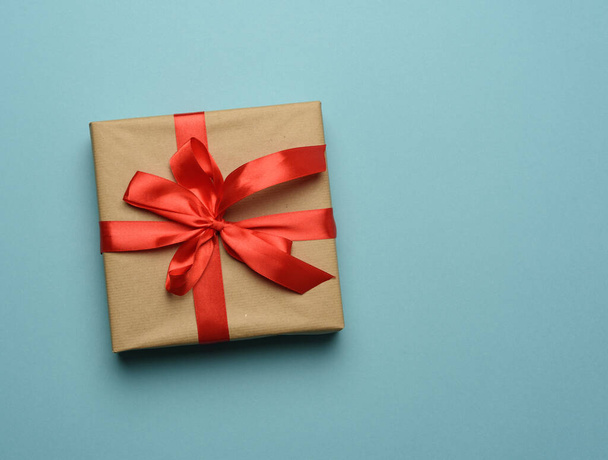 box wrapped in brown paper and tied with a red silk ribbon with a bow, gift on a blue background, top view, copy space - Valokuva, kuva