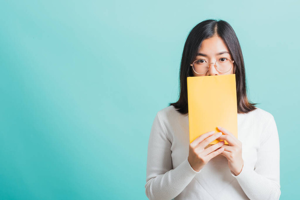 Young beautiful Asian woman hiding behind an open book, Portrait female in glasses is holding and reading a book, studio shot isolated on a blue background, Education concept - Zdjęcie, obraz
