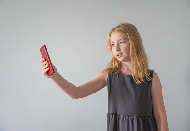 a girl in a gray dress is talking on a video link from a red mobile phone on gray background - Photo, Image