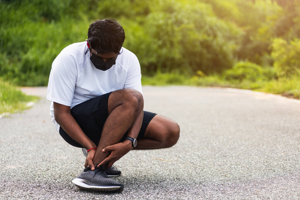 Close up Asian young sport runner black man wear watch hands joint hold leg pain because of twisted ankle broken while running at the outdoor street health park, healthy exercise Injury from workout - 写真・画像