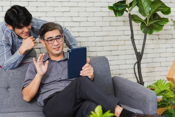 Asian senior businessman with digital tablet discuss together with young team in office. Father man and his son sit on sofa talking chatting on video call conference on tablet in living room at home - Foto, afbeelding