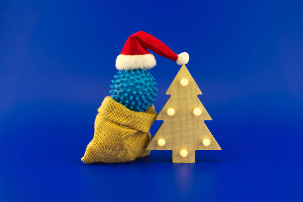 Christmas in pandemic concept still life with blue virus molecule, red Santa hat, Christmas tree and gift box sitting on face mask all isolated on blue background - Foto, Imagem