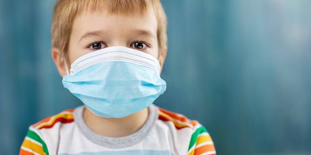 Boy in medical face protection mask indoors on blue background. Sick child. Coronavirus safety and allergy concept - Photo, Image