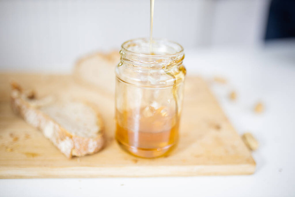 Honey jar and a slice of bread on wooden cutting board with blurry background. Stream of honey pouring into glass jar. Sweet and light breakfast - Photo, image