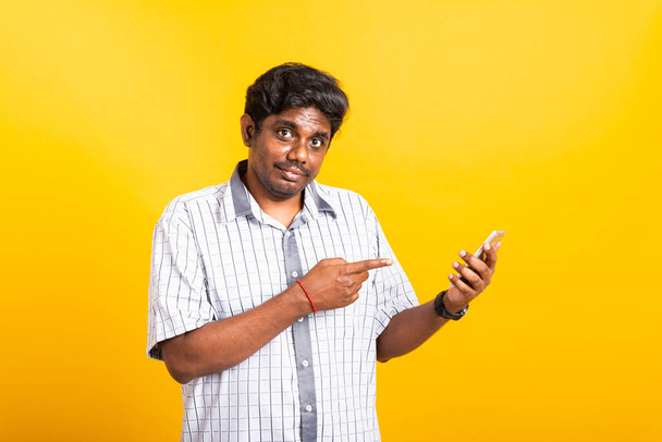 Asian happy portrait young black man smile standing wear shirt making finger pointing on smart digital mobile phone blank screen isolated, studio shot yellow background with copy space - 写真・画像