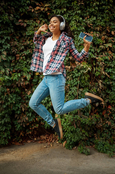 Happy woman in headphones jumping in summer park. Female music fan walking outdoors, girl in earphones, green bushes on background - Fotografie, Obrázek