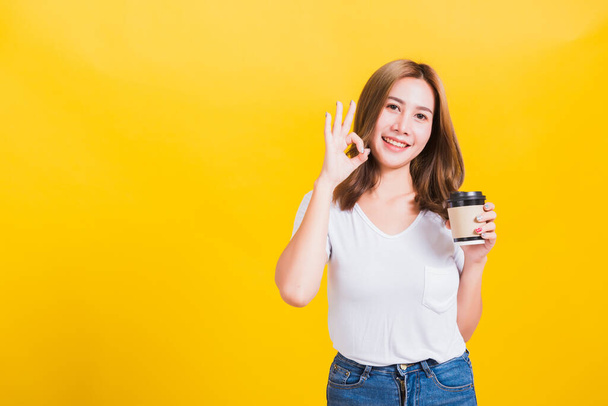 Portrait Asian Thai beautiful happy young woman standing holds take away cup coffee paper and confident show OK sign with fingers excellent, studio shot isolated on yellow background, with copy space - Foto, afbeelding