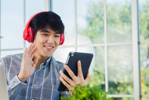 Asian young business man happiness with red headphones he sitting on desk workplace using digital tablet while greeting by video chat conferences talking and discussing - Photo, image