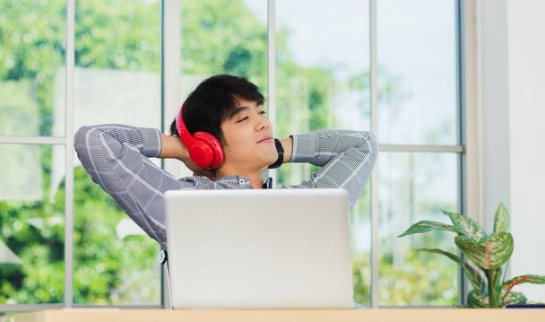 Asian young business man smile listening music in red headphone at home office. Happy businessman sitting on desk and relax wear wireless head phones enjoy listen music and work with laptop computer - Фото, зображення