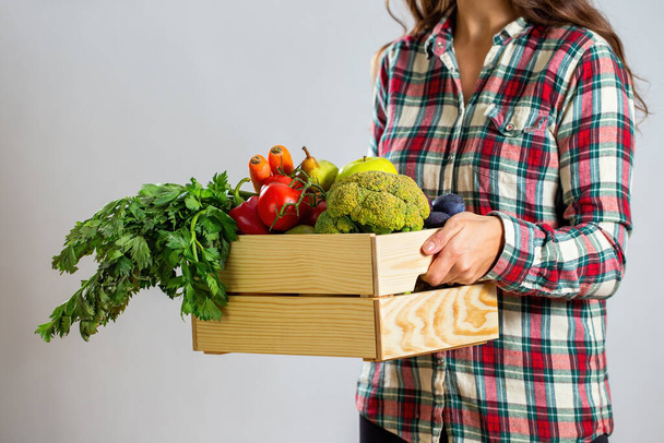 Woman holding box full of fruit and vegetable on white background. Wooden crate filled with fresh broccoli, tomatoes, carrot and celery. Lady in flannel checkered shirt shirt with healthy pack. - Photo, image