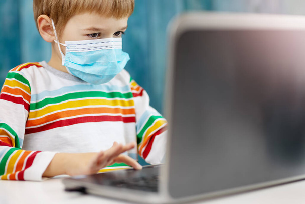Boy in medical face protection mask indoors on blue background. Child sitting at the table and studying on the laptop. - Foto, Imagem