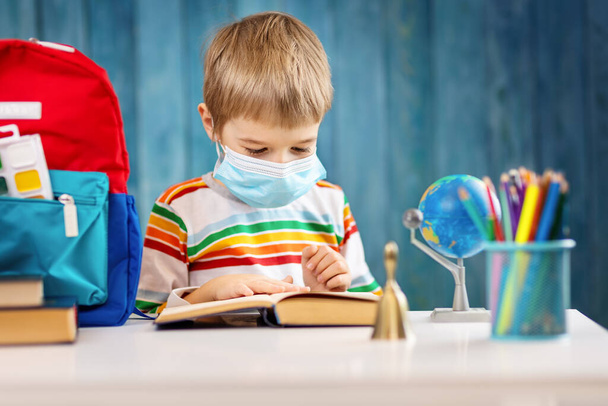 Boy in medical face protection mask indoors on blue background. Sick child sitting at the table and reading a book. Coronavirus pandemic. - Фото, зображення