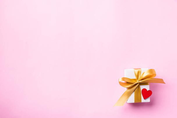 Valentines 'day background concept. Bílá dárková krabička se zlatou mašlí a dřevěnou červenou srdíčkovou kompozicí blahopřání pro šťastnou lásku izolovanou na růžovém pozadí s kopírovacím prostorem. Pohled shora - Fotografie, Obrázek