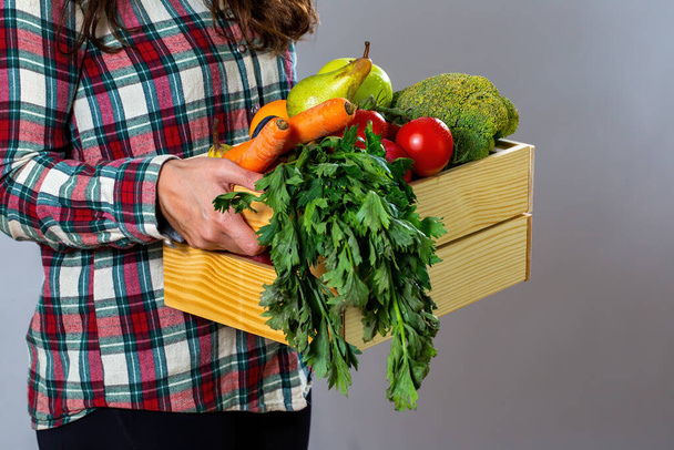 Person in flannel shirt holding box filled with fruit and vegetable. Woman with wooden crate full of fresh ripe food. Brunette showing healthy organic plants. - Photo, image