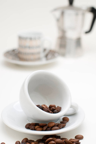 Tilted white espresso cup on white saucer with roasted coffee beans in, on and in front of the saucer in front of blurred cup with coffee pot on white background. - Foto, afbeelding