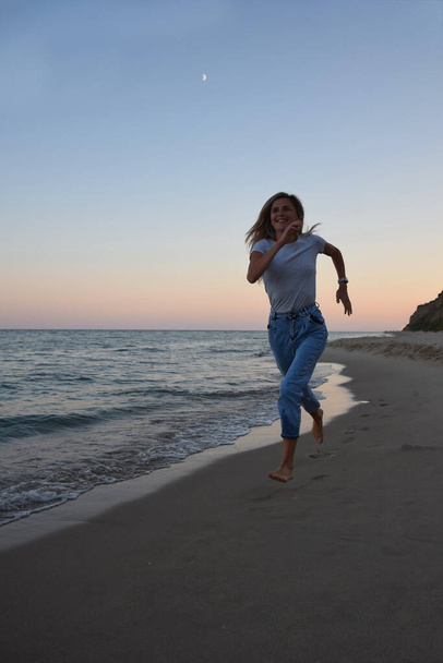 Una chica rubia con el pelo largo en jeans y una camiseta corre a lo largo de la orilla del mar de la noche. Vista frontal - Foto, imagen